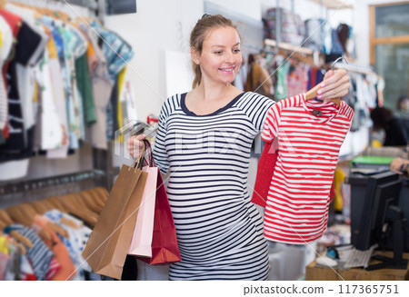 Pregnant woman in striped tunic chooses shirt in clothing shop Pregnant woman in striped tunic chooses shirt in clothing shop 117365751