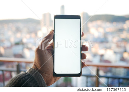 A Hand Gripping a Smartphone Set Against a Stunning Cityscape Background in Daylight A Hand Gripping a Smartphone Set Against a Stunning Cityscape Background in Daylight 117367513