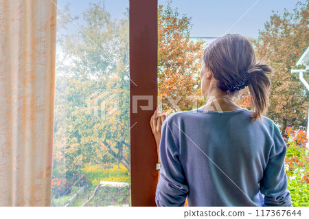 An unrecognizable woman on an open veranda looks out at the rain and yellowed autumn leaves in garden. Rear view An unrecognizable woman on an open veranda looks out at the rain and yellowed autumn leaves in garden. Rear view 117367644
