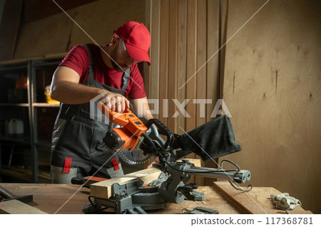 A woodworker is demonstrating the use of a miter saw to achieve accurate cuts on various pieces of lumber. The workspace is well-organized, featuring tools and equipment essential for woodworking 117368781