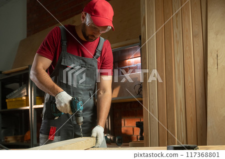 A skilled craftsman utilizes a drill to construct wooden projects in a well-organized workshop. The warm light sets the perfect atmosphere for detailed work, showcasing his focus and expertise. A skilled craftsman utilizes a drill to construct wooden projects in a well-organized workshop. The warm light sets the perfect atmosphere for detailed work, showcasing his focus and expertise. 117368801