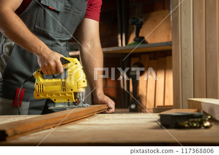 A skilled craftsman is focused on his woodworking project, using a electric jigsaw to cut timber precisely. The workshop is well-organized, with various tools and materials surrounding him, showcasing A skilled craftsman is focused on his woodworking project, using a electric jigsaw to cut timber precisely. The workshop is well-organized, with various tools and materials surrounding him, showcasing 117368806