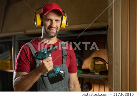 A man in a red cap and overalls puts on hearing protection equipment in the workshop. The room is filled with various tools and equipment, which indicates active woodworking work. 117368810