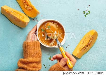 Woman holding spoon eating butternut squash soup with cream on blue background top view 117369004