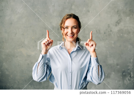 Cheerful young businesswoman in a blue shirt points upward, offering a welcoming and positive Cheerful young businesswoman in a blue shirt points upward, offering a welcoming and positive 117369056