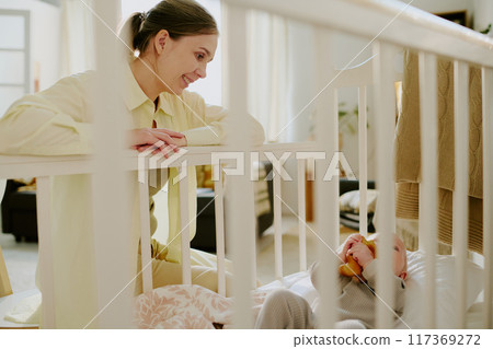 Smiling mother engaging with baby in crib holding a stuffed toy. Room decorated with warm, natural tones with calming atmosphere enhancing bond between parent and child 117369272