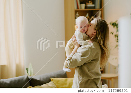 Loving mother holding baby while standing in cozy living room with soft natural light creating warm atmosphere adorable child snuggling close and feeling secure 117369291