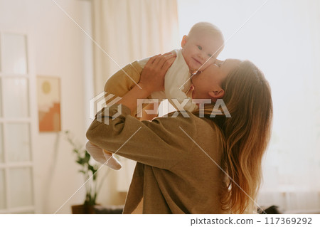 Mother holding her baby in cozy living room setting with warm natural light, creating intimate and loving moment, soft, nurturing environment with indoor plant and framed painting visible Mother holding her baby in cozy living room setting with warm natural light, creating intimate and loving moment, soft, nurturing environment with indoor plant and framed painting visible 117369292
