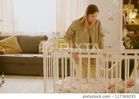 Mother standing by crib watching her sleeping baby. Soft beige decor and sunlight from large windows create a warm and serene ambiance in living room Mother standing by crib watching her sleeping baby. Soft beige decor and sunlight from large windows create a warm and serene ambiance in living room 117369293