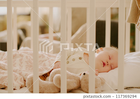 A peaceful baby is sleeping soundly in a cozy crib while holding a fluffy teddy bear and surrounded by a warm blanket A peaceful baby is sleeping soundly in a cozy crib while holding a fluffy teddy bear and surrounded by a warm blanket 117369294