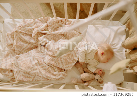 Peaceful scene of baby sleeping in crib cuddling with stuffed animal and covered by soft blanket surrounded by soothing environment enhancing serenity Peaceful scene of baby sleeping in crib cuddling with stuffed animal and covered by soft blanket surrounded by soothing environment enhancing serenity 117369295