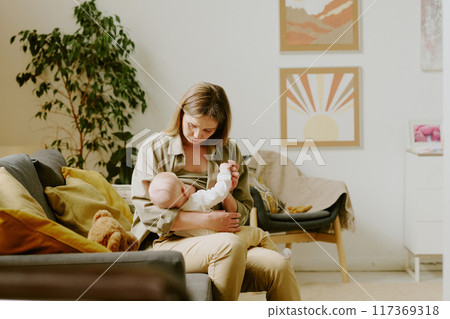 Mother holding and feeding baby while sitting on couch in cozy living room with sun painting and greenery. Painting depicts abstract sun setting behind mountains 117369318