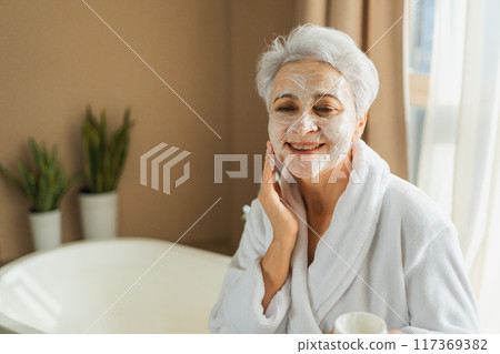 Mature adult old woman applying nourishing anti age face cream doing daily beauty routine. Senior woman with white mask or creme on skin gets facial treatment Skin care aging beauty skincare cosmetics Mature adult old woman applying nourishing anti age face cream doing daily beauty routine. Senior woman with white mask or creme on skin gets facial treatment Skin care aging beauty skincare cosmetics 117369382