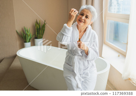 Woman applying natural drops cosmetics oil to face skin. Mature adult old woman applying nourishing anti age serum with pipette doing daily beauty routine. Senior woman gets kin scare facial treatment 117369388
