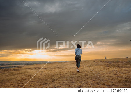 A person is running in an open field during sunset with dramatic clouds in the sky 117369411