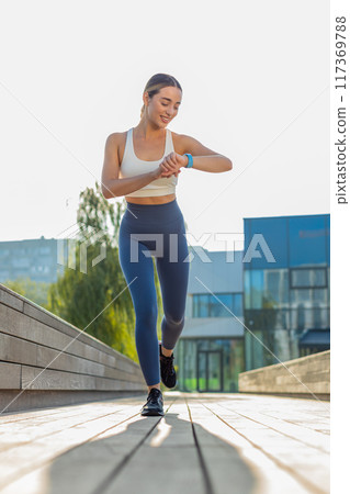 Happy athletic runner adult girl looks at smartwatch device fitness bracelet checking time in park 117369788