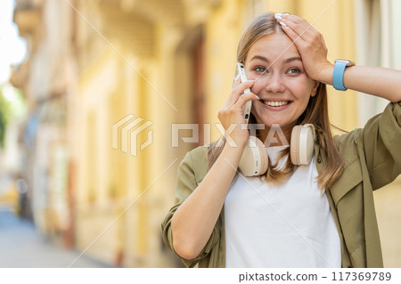 Happy surprised Caucasian young woman in pleasant conversation on smartphone standing on city street 117369789