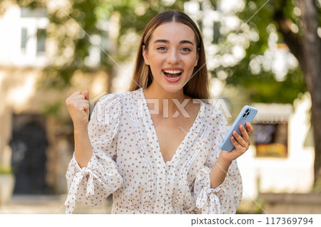 Excited happy woman using smartphone celebrating win receive good message news in urban street 117369794