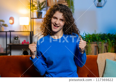 Happy woman celebrating success showing thumbs up dancing sits on sofa in living room at home 117369822