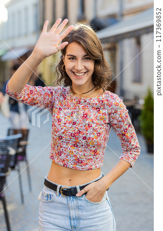 Young woman smiling friendly at camera, waving hands hello, hi, greeting or goodbye in city street Young woman smiling friendly at camera, waving hands hello, hi, greeting or goodbye in city street 117369852