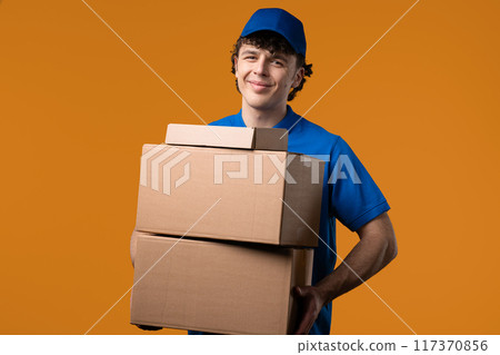 Positive postal courier man with parcel box. Happy postman delivers address package on yellow studio background. 117370856