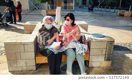 Mother and Daughter Rest After Flu Shots on Sunny Bench Outside Medical Center, Wearing Masks and Reflecting on Health and Safety Protocols. 117372149