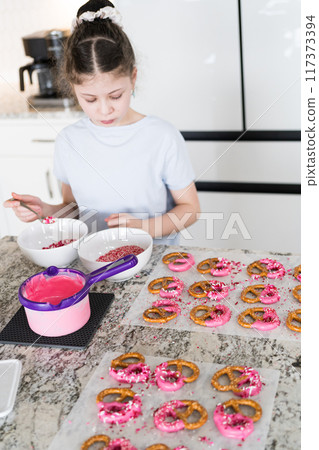 With focused attention, this budding culinary artist dips pretzels into a pot of melted chocolate, creating sweet delights in the warmth of a well-lit home kitchen. 117373394