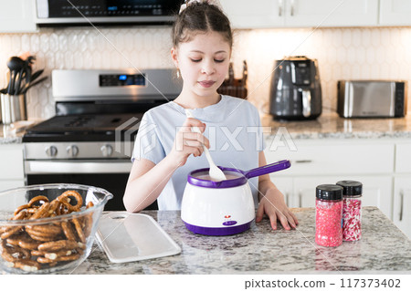 With focused attention, this budding culinary artist dips pretzels into a pot of melted chocolate, creating sweet delights in the warmth of a well-lit home kitchen. With focused attention, this budding culinary artist dips pretzels into a pot of melted chocolate, creating sweet delights in the warmth of a well-lit home kitchen. 117373402