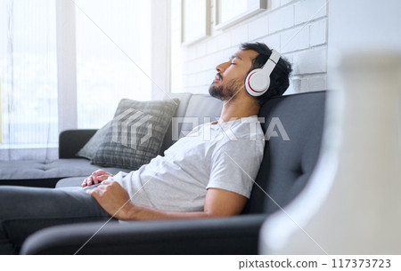 Music, relax and man with headphones on sofa in home living room streaming radio or podcast. Meditation, technology and male on couch in lounge listening to peaceful song, audio or album in house. Music, relax and man with headphones on sofa in home living room streaming radio or podcast. Meditation, technology and male on couch in lounge listening to peaceful song, audio or album in house. 117373723