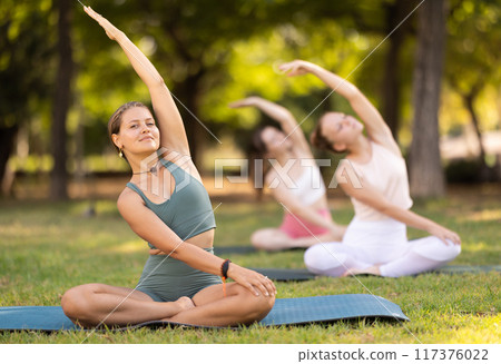 Portrait of women sitting on gym mat and doing yoga meditation in lotus position and arms raised in park 117376022