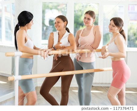 Young ballerinas chatting joyfully standing around training barre 117376471