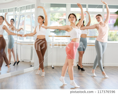Young female ballet dancer executing dance moves with focused precision among group of people rehearsing in ballet studio 117376596