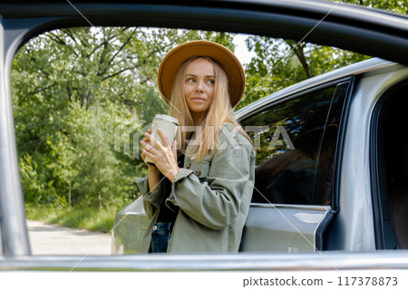 Blonde woman stoped on road next to car and drink coffee or tea from reusable mug. Refuse reuse recycle zero waste concept. Young tourist explore local travel making candid real moments. Responsible 117378873