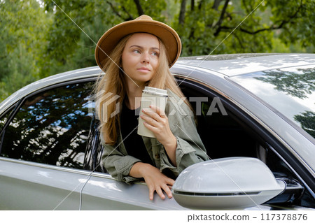 Smiling young woman looking from car window and drinking coffee or tea from reusable thermos cup. Local solo travel on weekends concept. Exited woman explore freedom outdoors in forest. Unity with Smiling young woman looking from car window and drinking coffee or tea from reusable thermos cup. Local solo travel on weekends concept. Exited woman explore freedom outdoors in forest. Unity with 117378876