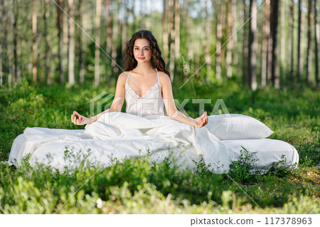 Young woman is sitting in the forest on a white bed Young woman is sitting in the forest on a white bed 117378963