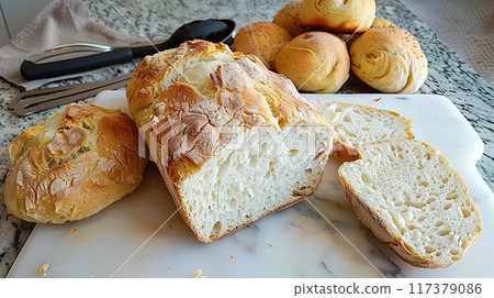 A loaf of freshly baked artisan bread sliced open, showcasing its airy texture, placed on a cutting board with rolls in the background.. A loaf of freshly baked artisan bread sliced open, showcasing its airy texture, placed on a cutting board with rolls in the background.. 117379086
