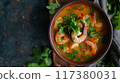 A flavorful bowl of spicy shrimp soup garnished with fresh cilantro and chili peppers, served with lime wedges on the side.. 117380031