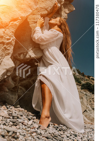 Woman summer travel sea. Happy lady with long hair in white dress enjoy taking photo outdoors for memories. Woman traveler posing on beach at sea surrounded by volcanic mountains, sharing emotions 117380478