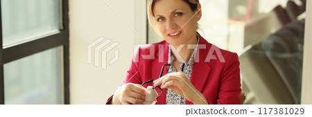 Smiling woman sitting in office and wiping glasses Smiling woman sitting in office and wiping glasses 117381029