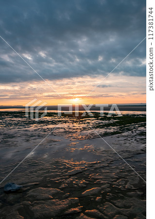 Tranquil sunset at a low tide beach with reflective waters under a cloudy sky, serene landscape Tranquil sunset at a low tide beach with reflective waters under a cloudy sky, serene landscape 117381744