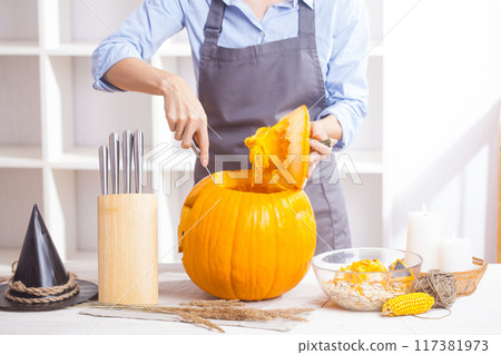 Woman in apron cuting out Halloween pumpkin, at wooden table, preparing holiday decorations at home 117381973