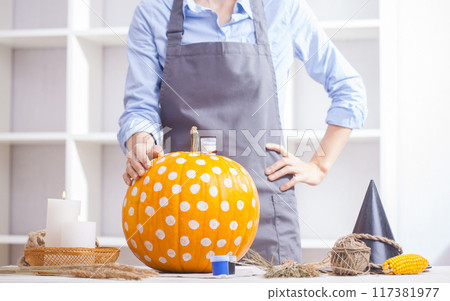 Woman in apron painting Halloween pumpkin, at wooden table, preparing holiday decorations at home 117381977