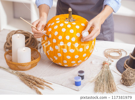 Woman in apron painting Halloween pumpkin, at wooden table, preparing holiday decorations at home 117381979
