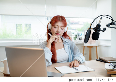 Attractive Asian female podcaster or radio journalist with red curly hair is working in her studio 117382127