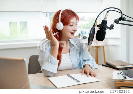 Beautiful Asian female podcaster with red curly hair is working in her studio, speaking into the mic Beautiful Asian female podcaster with red curly hair is working in her studio, speaking into the mic 117382131