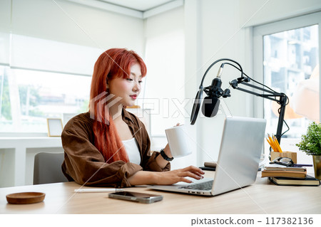 An attractive Asian female podcaster is working in her podcast studio, drinking coffee. 117382136