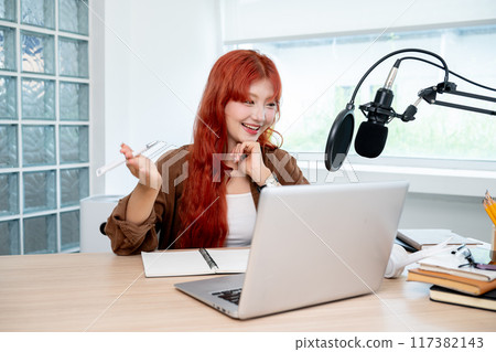 A smiling Asian female radio host or podcaster is working in her studio, speaking into the mic. A smiling Asian female radio host or podcaster is working in her studio, speaking into the mic. 117382143
