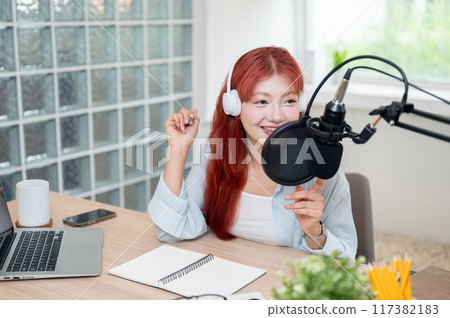 A beautiful Asian female podcaster is recording her podcast in the studio, speaking into a mic. A beautiful Asian female podcaster is recording her podcast in the studio, speaking into a mic. 117382183