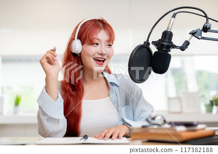 A beautiful Asian female podcaster is recording her podcast in the studio, speaking into a mic. A beautiful Asian female podcaster is recording her podcast in the studio, speaking into a mic. 117382188