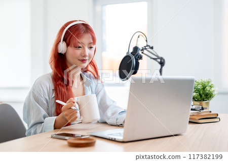 A beautiful Asian female podcaster is working in her home studio and enjoying a coffee. A beautiful Asian female podcaster is working in her home studio and enjoying a coffee. 117382199
